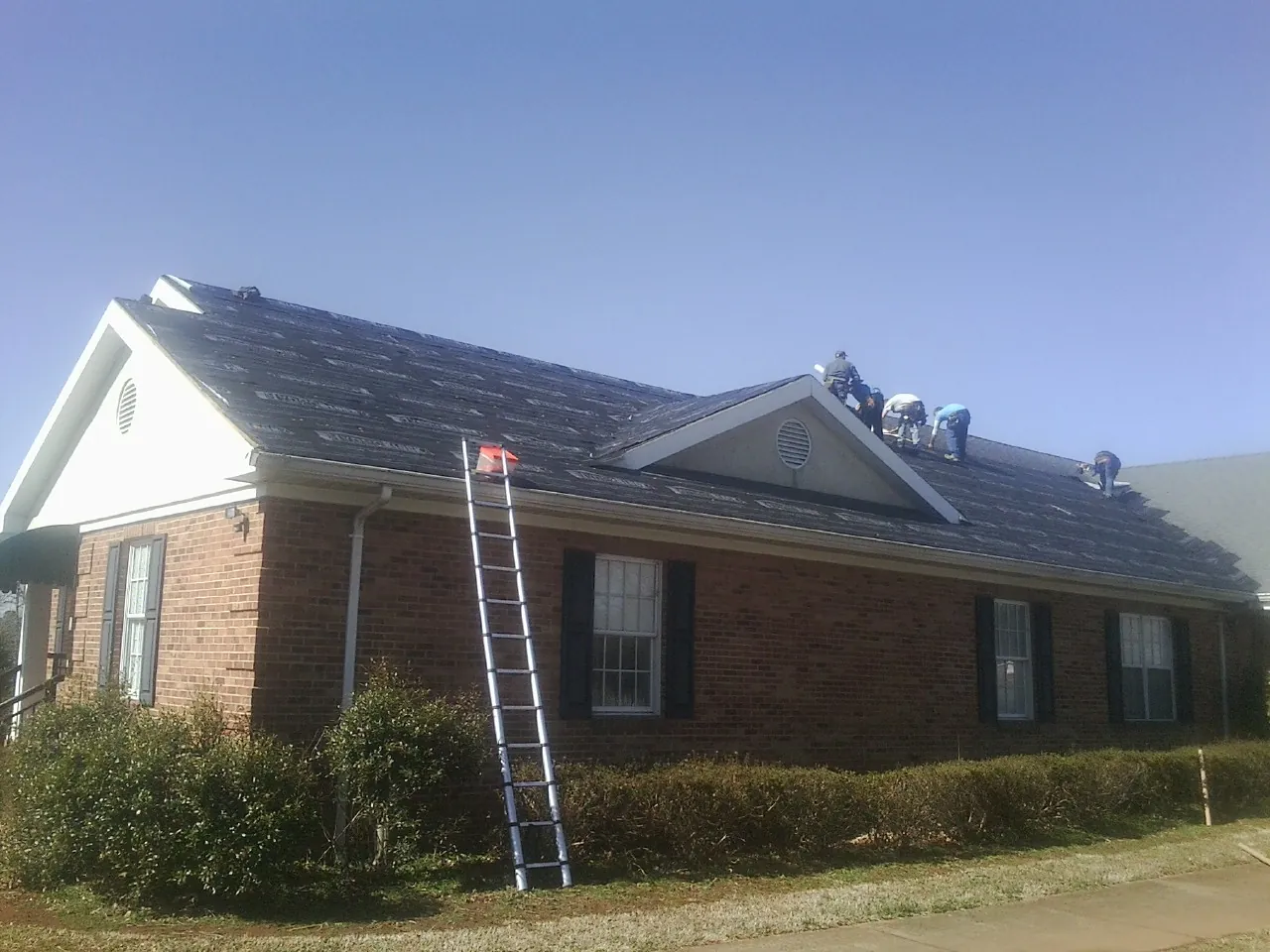 Crew tearing shingles off a brick ranch roof with a ladder set — Emergency Roof Repair work by our crew in Lovelady