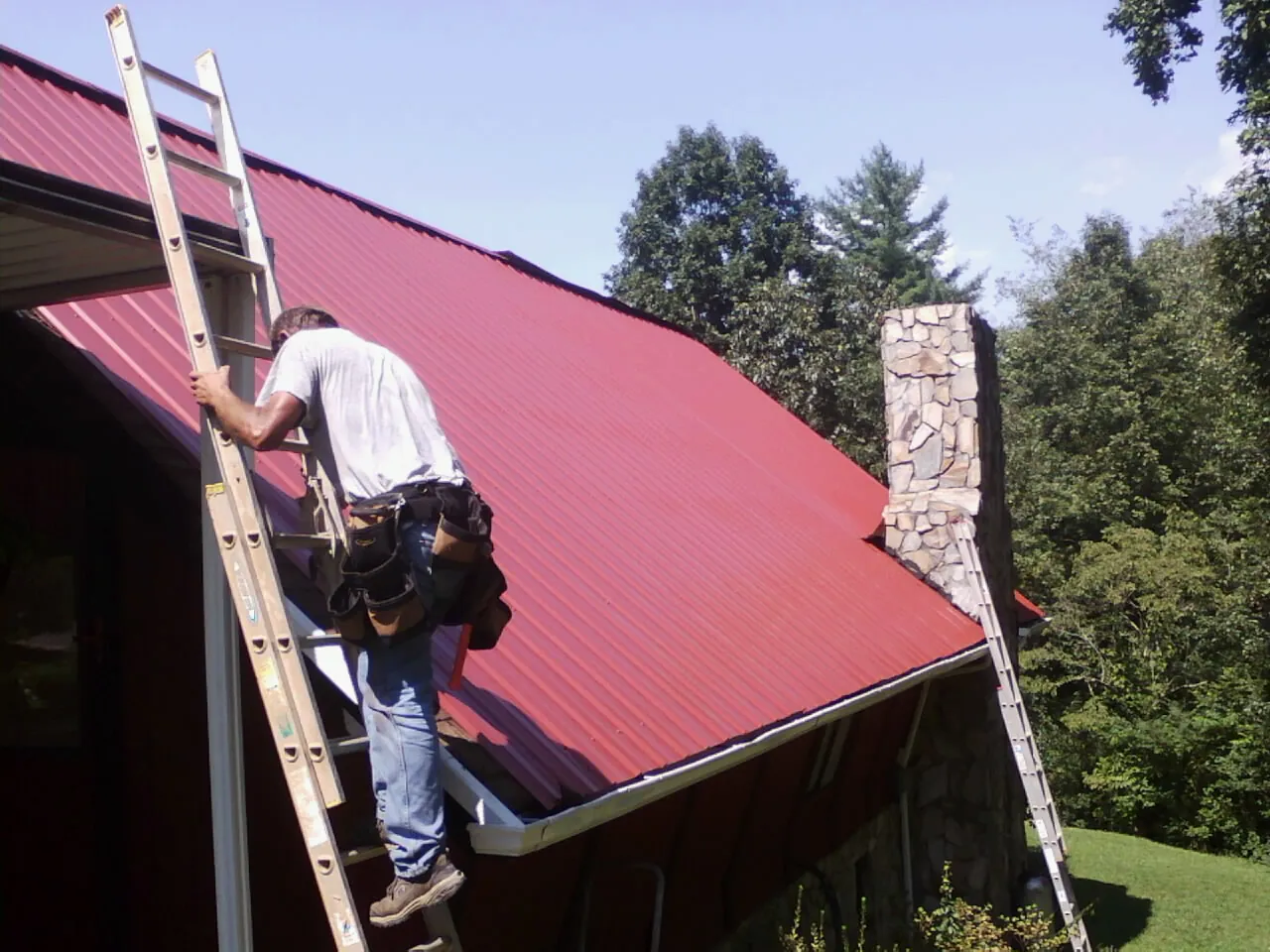 Roofer on a ladder beside a red metal roof and stone chimney — Soffit & Fascia Repair by Roofing Brothers in Lovelady