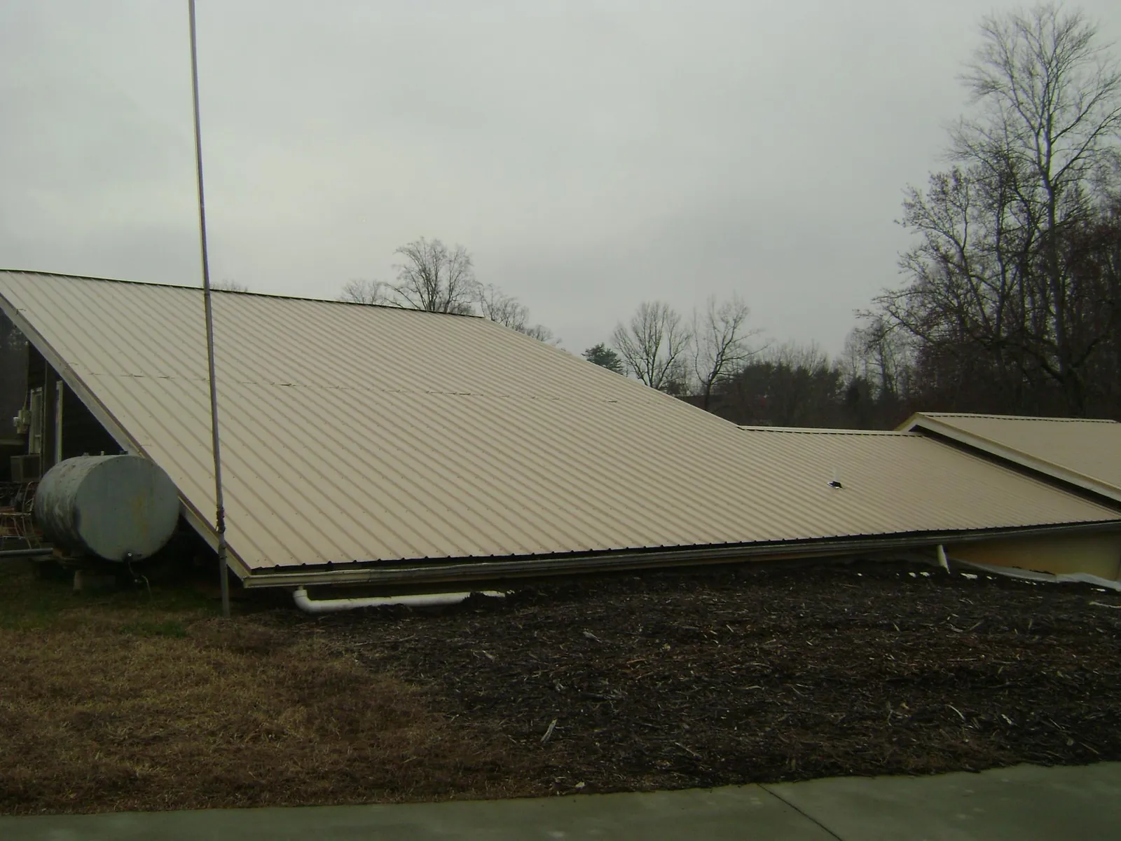 Wide tan metal roof plane seen from the ground beside a propane tank in Lovelady